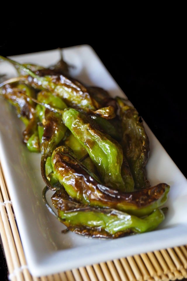 Blistered shishito peppers on a narrow white plate on a sushi mat.