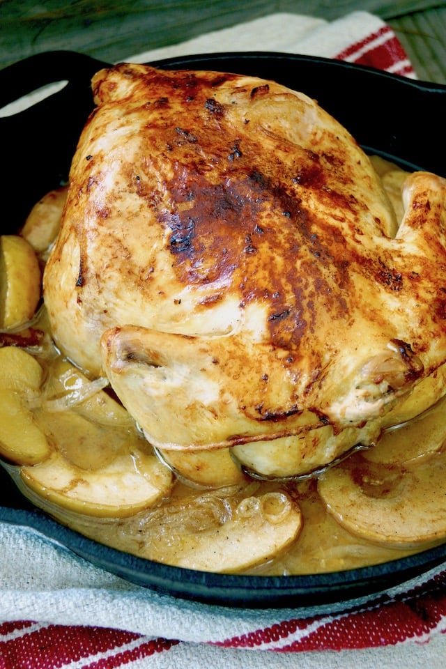 Whole roasted chicken surrounded by apples slices in a cast iron skillet.