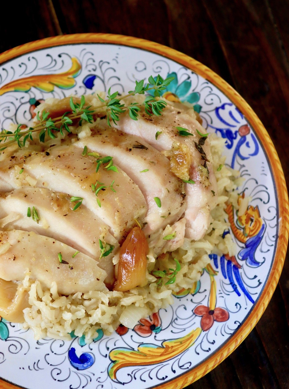 Sliced chicken thigh on brown rice with whole roasted garlic cloves, on Italian gold and blue painted plate.
