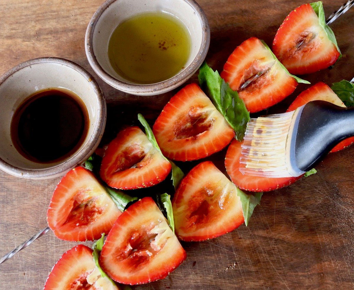 Two skewers of strawberries cut in half with basil leaves between them, being brushed with olive oil and balsamic vinegar.