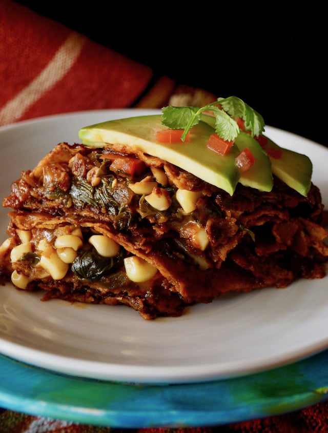 One vegetarian enchilada on a white plate, sliced in half so the filling is showing, with avocado slices and diced tomato on top.