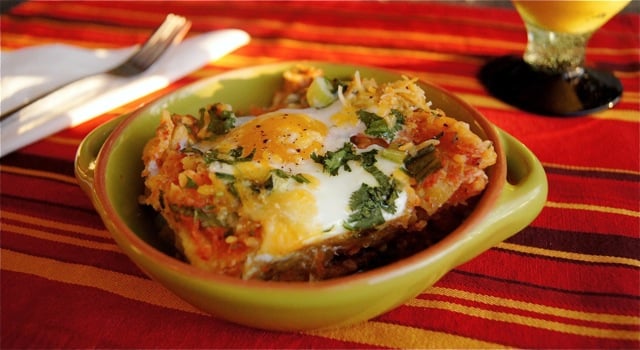 Smoky Chipotle Breakfast Nachos with Eggs in a light green, ceramic bowl on a red, gold and brown striped cloth, with glass of oj in background.