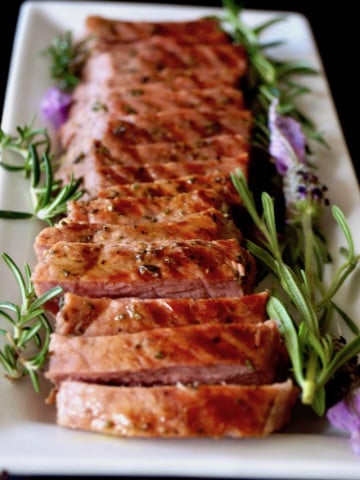 Sliced Lavender-Rosemary Grilled Ribeye Steak on a narrow white platter, showing grill marks, with fresh herbs.