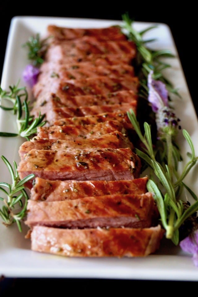 Sliced Lavender-Rosemary Grilled Ribeye Steak on a narrow white platter, showing grill marks, with fresh herbs.