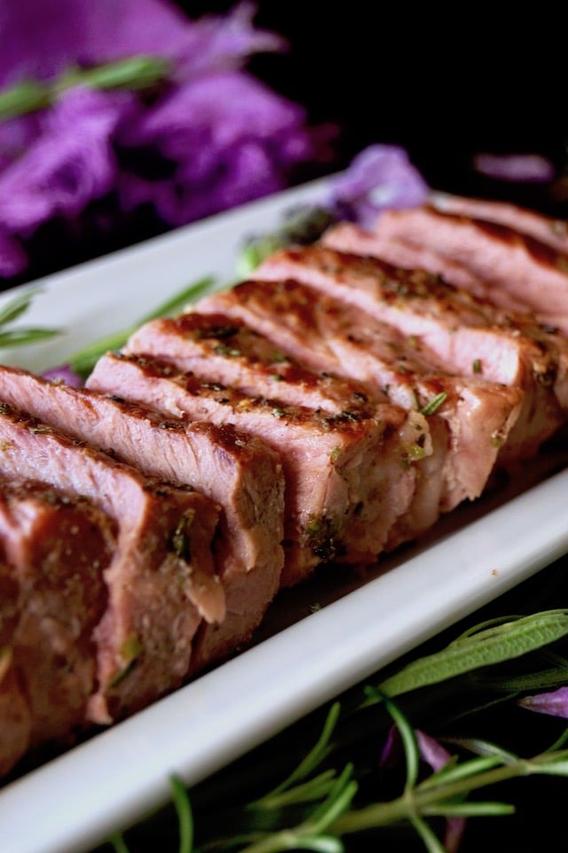 Close up of sliced Lavender-Rosemary Grilled Ribeye Steak on a narrow white platter with fresh herbs.