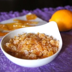 Candied Citrus Peels in a small white bowl on a purple cloth with 2 orange Meyer lemons in the background.