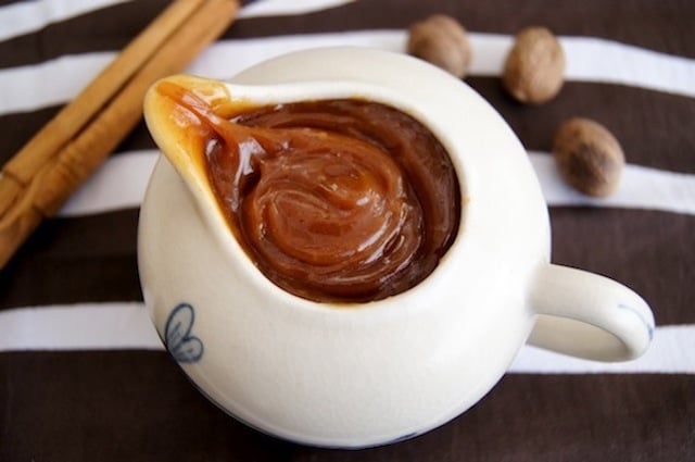 Spiced Caramel in a small white pitcher surrounded by a cinnamon stick and three whole nutmeg.