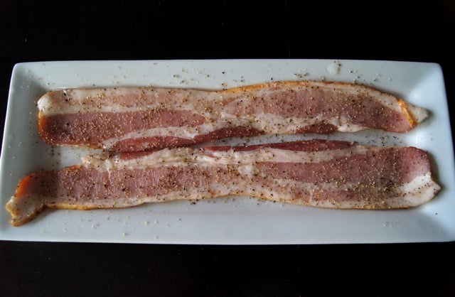 Two strips of raw bacon on a white plate with pepper, for Bloody Mary with Bacon.