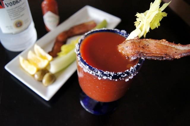 Bloody Mary with Bacon and celery in a glass with a blue rim coated in salt, with white plate of garnishes and vodka bottle in background.