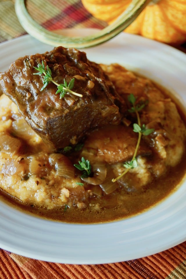 Serving of Pumpkin Braised Short Ribs on polenta with fresh thyme, in a white plate with pumpkin in background.