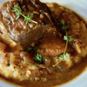 One serving of Pumpkin Braised Short Ribs on a bed of polenta with fresh thyme, in a white plate.