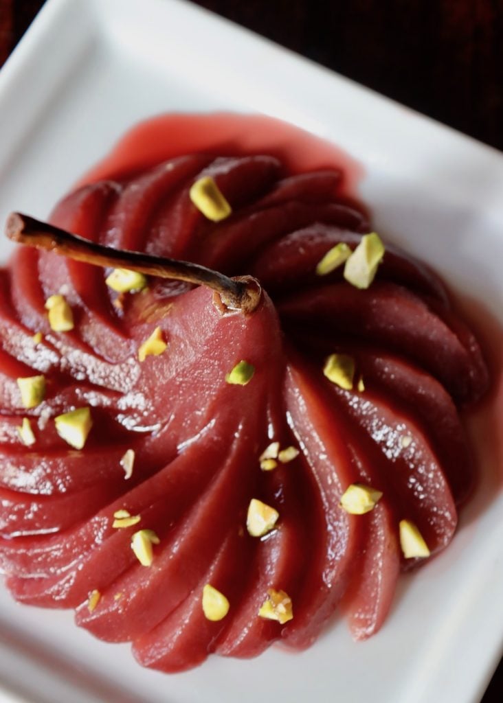 One whole fanned port wine poached pears on white dish with pistachios.