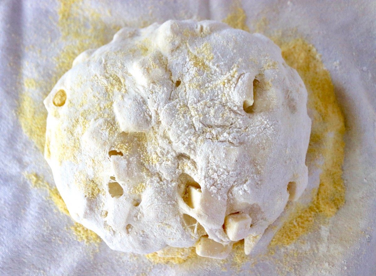 Evenly shaped round ball of Asiago bread dough on cornmeal on a tea towel.