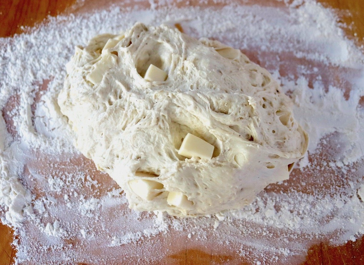 Misshapen ball of bread dough with chunks of cheese, on a floured wood surface.