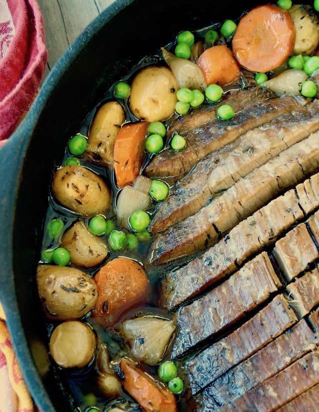 Sliced brisket on a cast iron skillet surrounded by potatoes, peas and carrots.