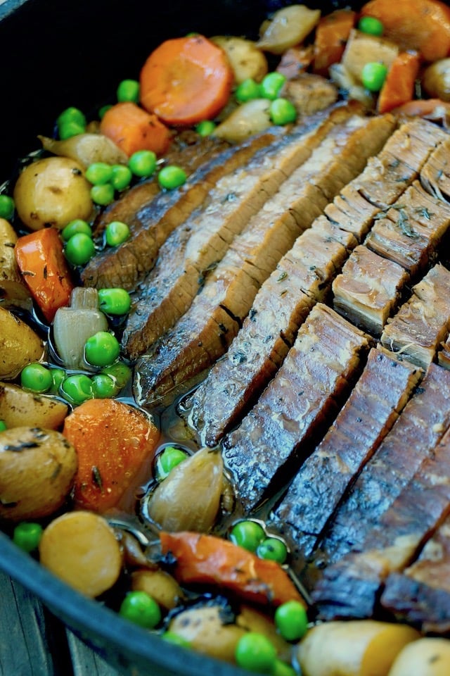 Close up of sliced brisket on a cast iron skillet surrounded by potatoes, peas and carrots.