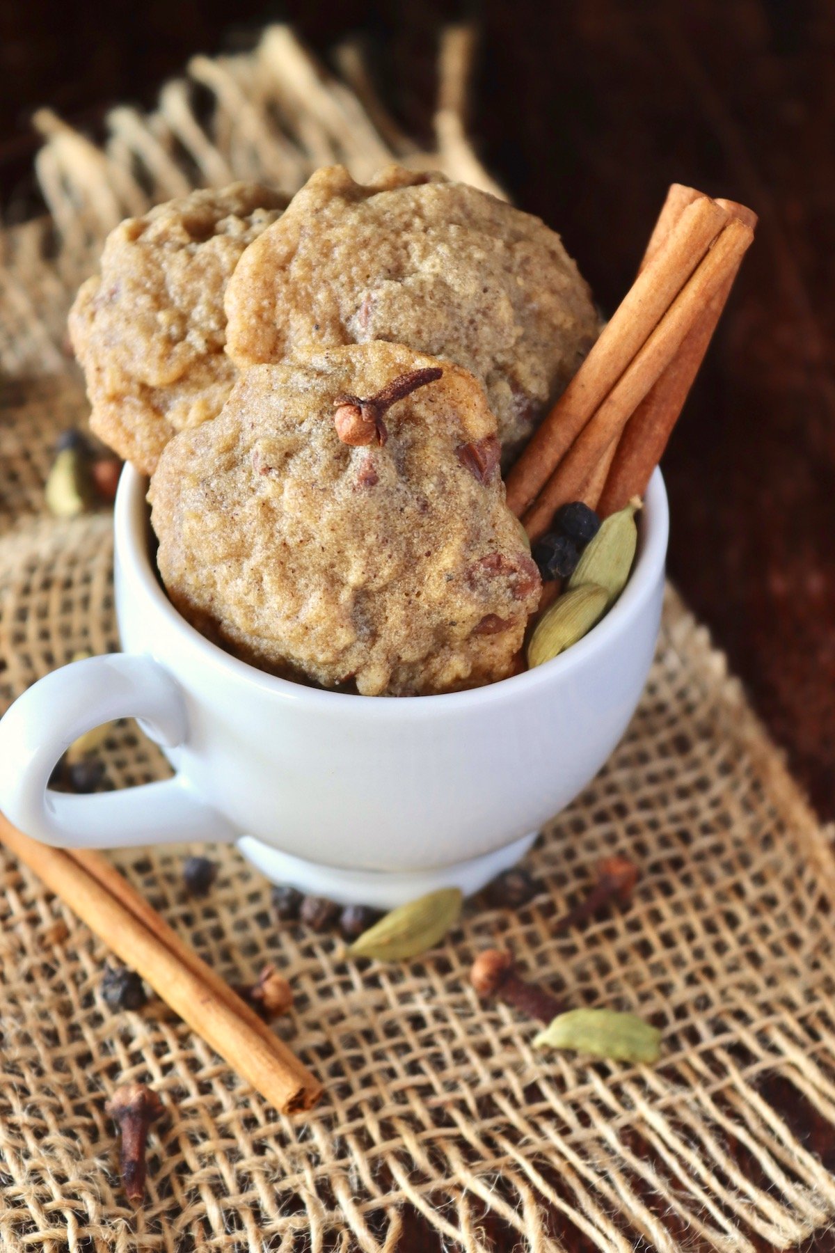 Small white espresso mugh holding 3 chai cookies with cinnamon sticks and green cardamom pods.