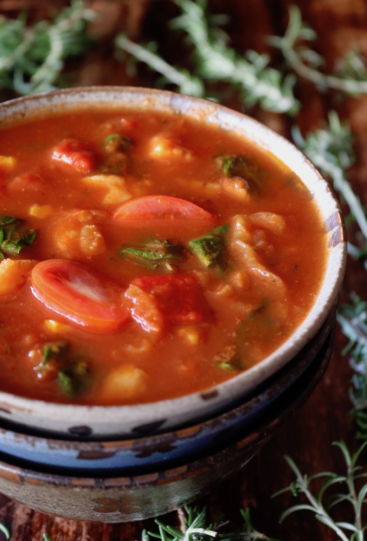 Three ceramic stacked bowl with the top one full of tomato fish stew, surrounded by rosemary