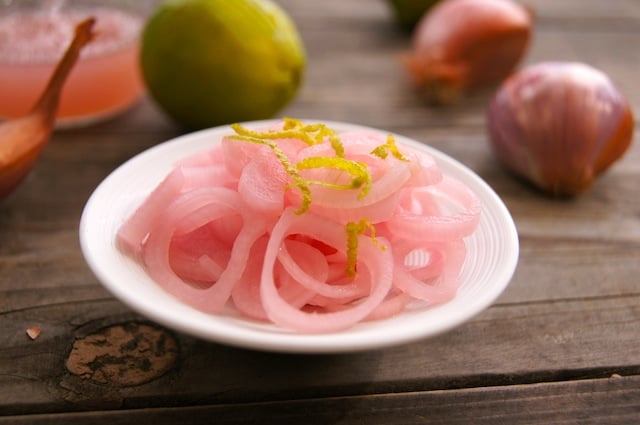 Pickled Shallots in a white bowl on wood table with while shallots and limes in background.