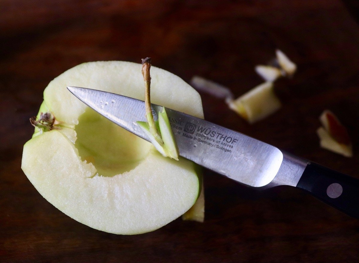 Half of a green apple with a paring knife slicing the stem off.