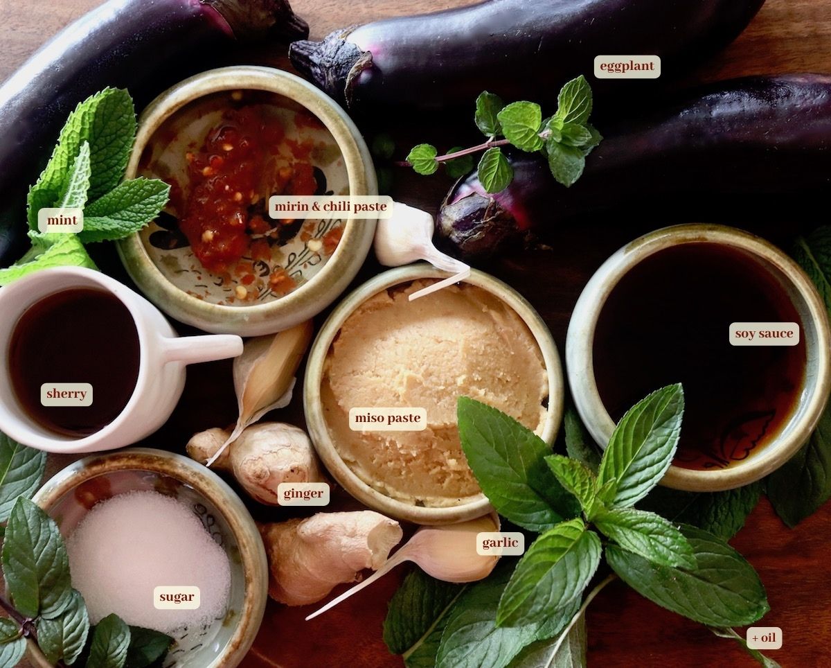 Ingredients for miso marinated eggplant in bowls and on cutting board, including miso paste, mint, soy sauce, ginger, garlic, chili paste, mirin, sugar and eggplant.