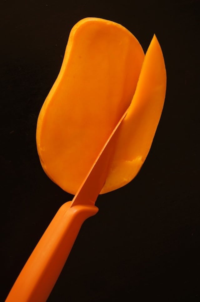Mango being sliced into a wedge off of the pit, with an orange knife