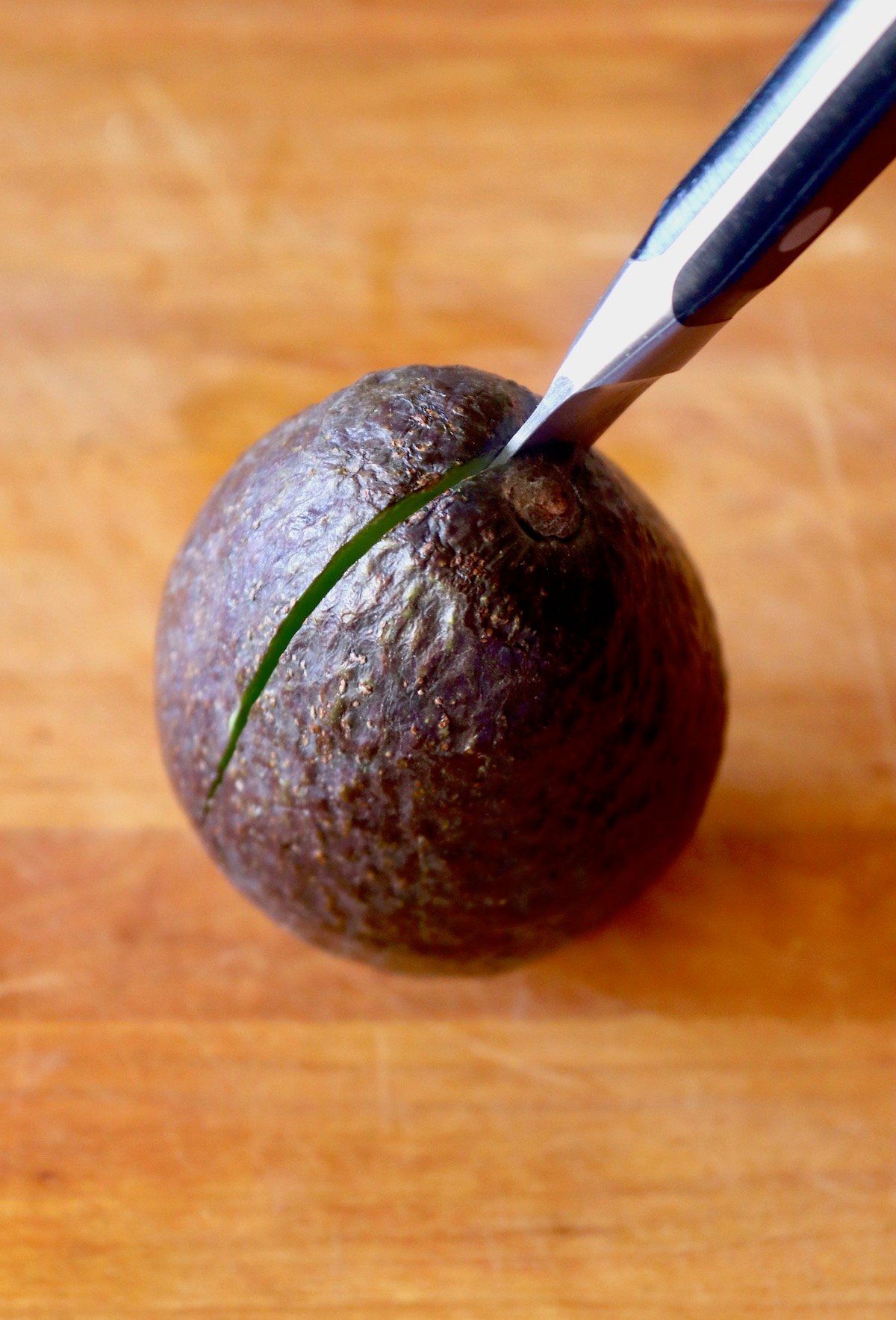 Avocado with a paring knife slicing it in half, starting at the tip, on wood cutting board.