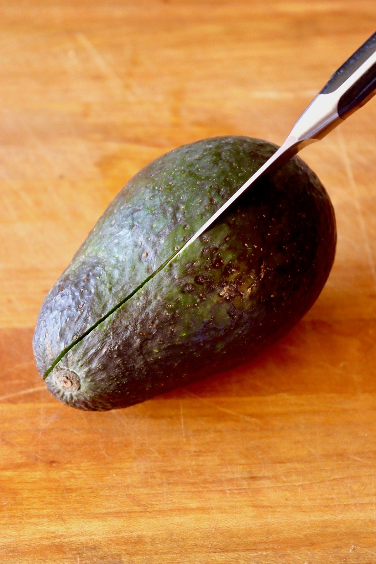 Avocado with a paring knife slicing it in half on wood cutting board.