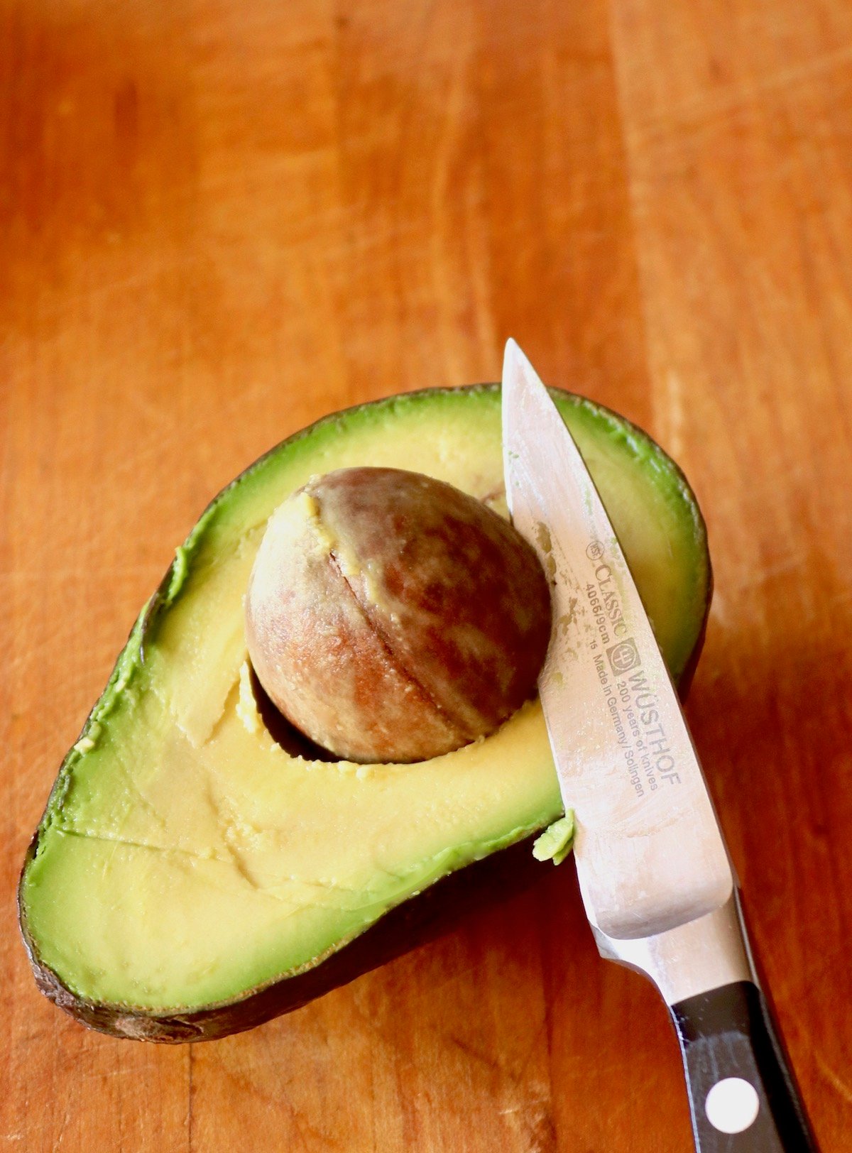 Avocado half on wood board with the pit in place with a paring knife cutting into it and twisting it a bit.