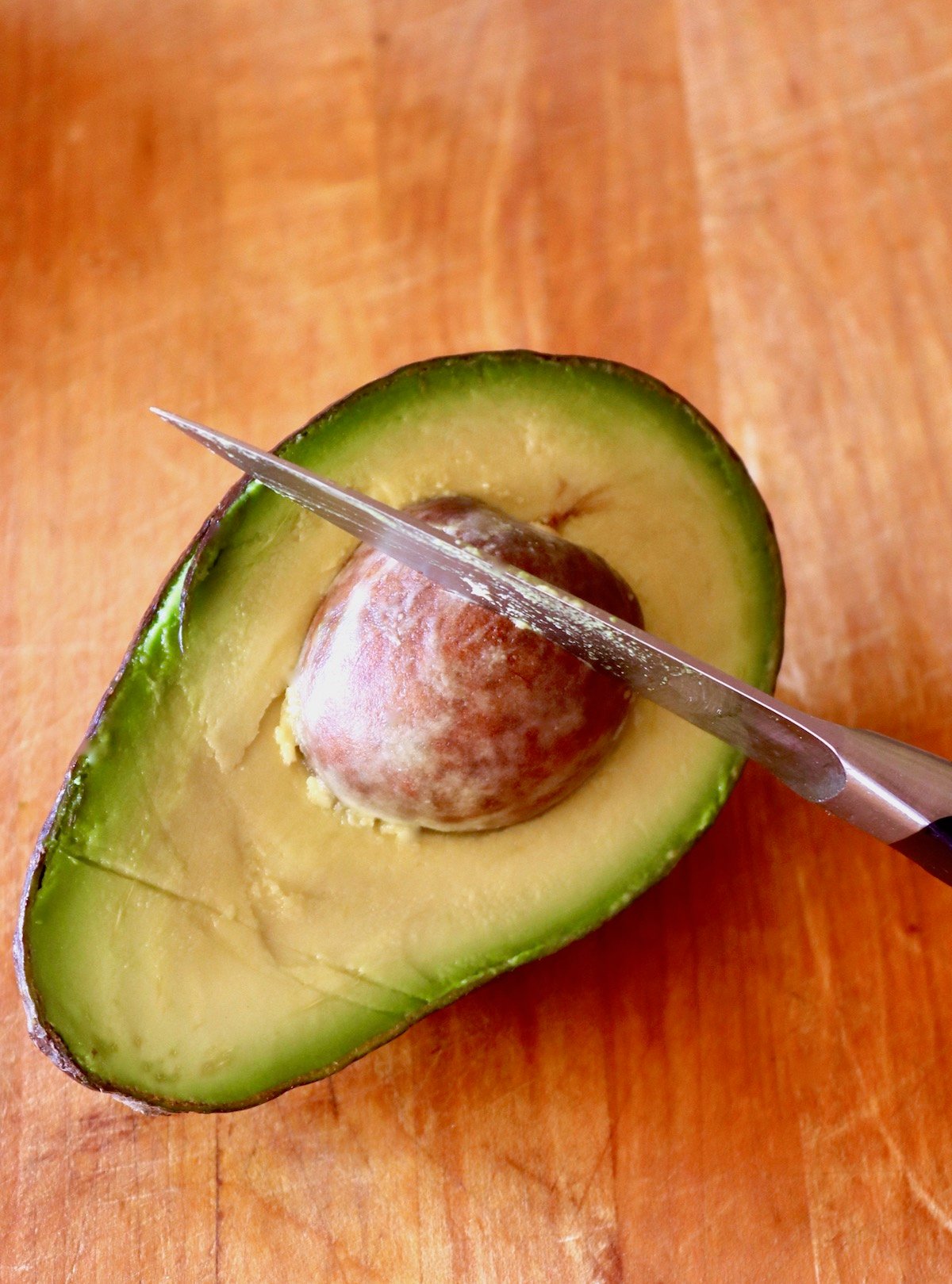 Avocado half on wood board with the pit in place with a paring knife cutting into it.