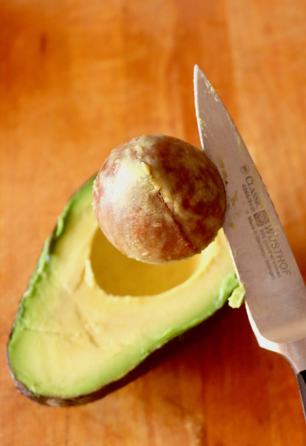 Avocado half on wood board with the pit in the air above it, with a paring knife stuck to it.