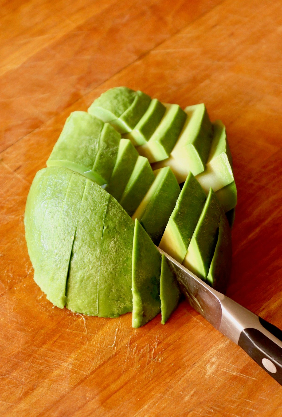 Peeled avocado, round side up on a wooden cutting board, being cut into a large dice.