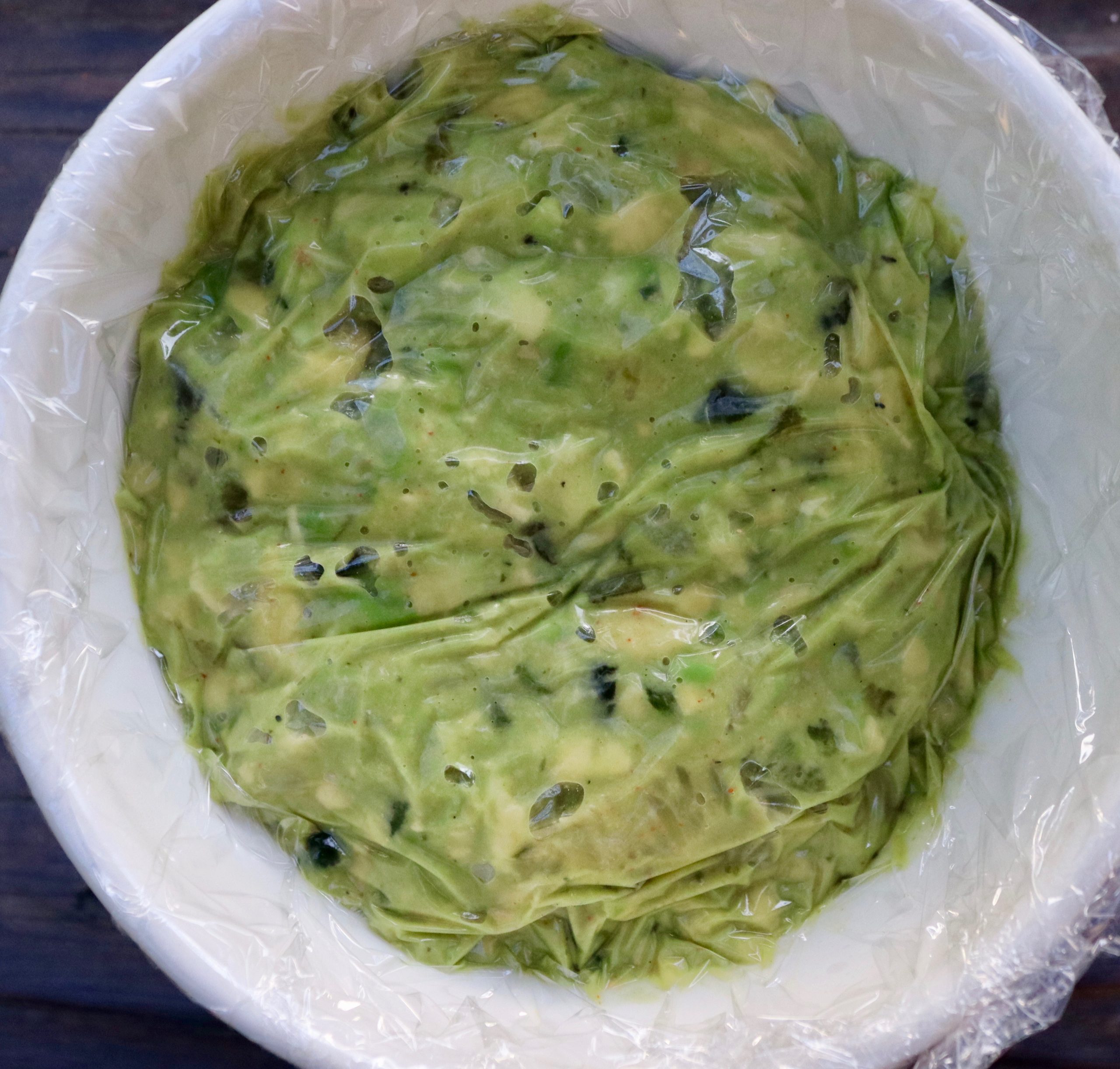 White bowl full with guacamole, covered with plastic wrip.