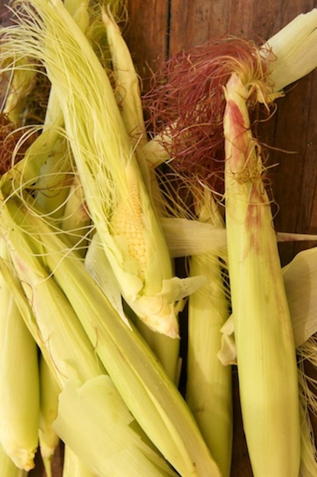 Baby corn in husks with purple silk showing, on wood surface.