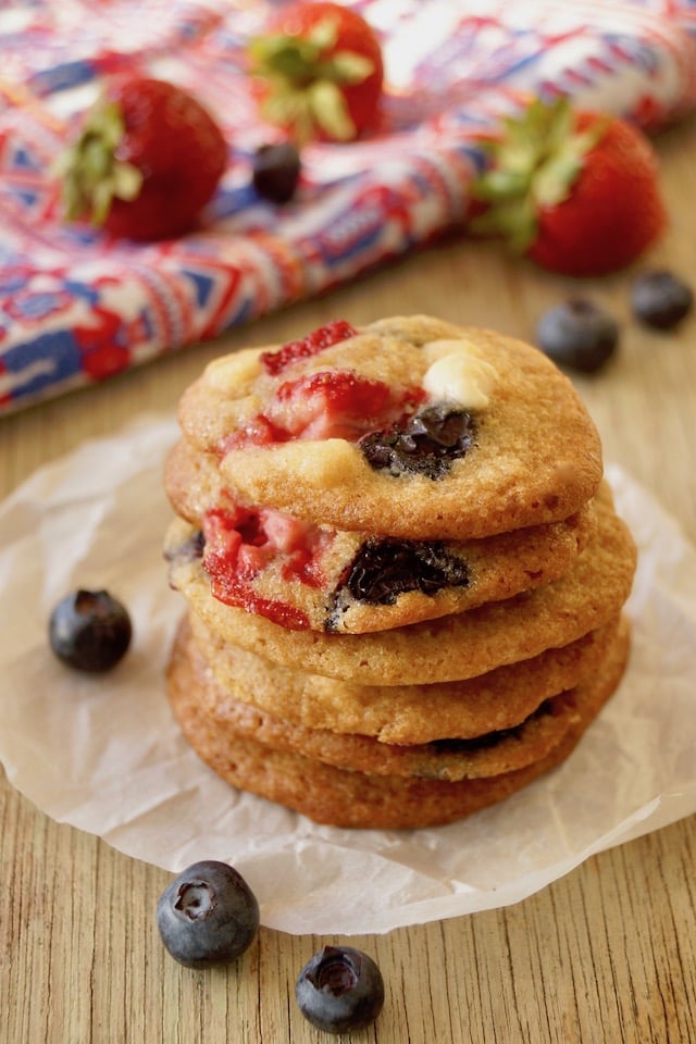 Stack of 6 Strawberry-Blueberry White Chocolate Chip cookies with a few blueberries and strawberries in background.