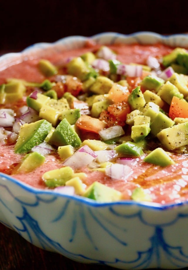 Tomato Gazpacho recipe in a blue and white bowl with diced aocados, tomatoes and onion on top