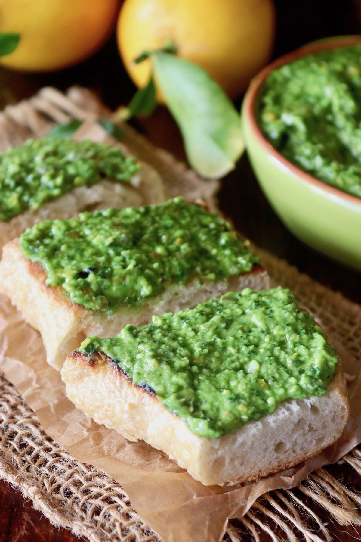Three small pieces of French bread with a thick spread of pesto on top, on crinkled parchment paper with lemons behind them.