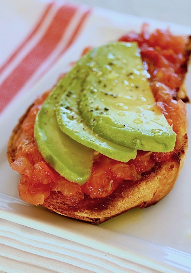Toast with breakfast tomatoes and avocado slices on a white plate.