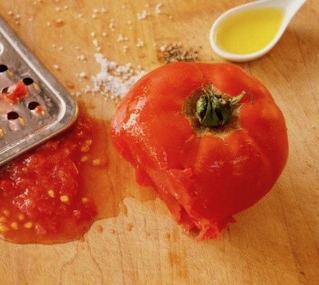 Cutting board with salt, pepper, olive oil land a partially grated tomato.