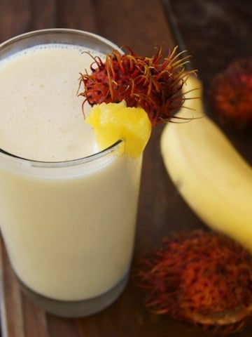 Tall glass filled with a tropical smoothie with a banana and rambutans in the background.