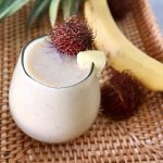 Rounded glass filled pale yellow-colored smoothie with a rambutan on the edge.
