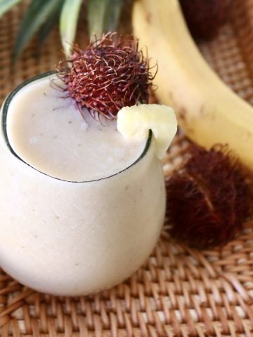Rounded glass filled pale yellow-colored smoothie with a rambutan on the edge.