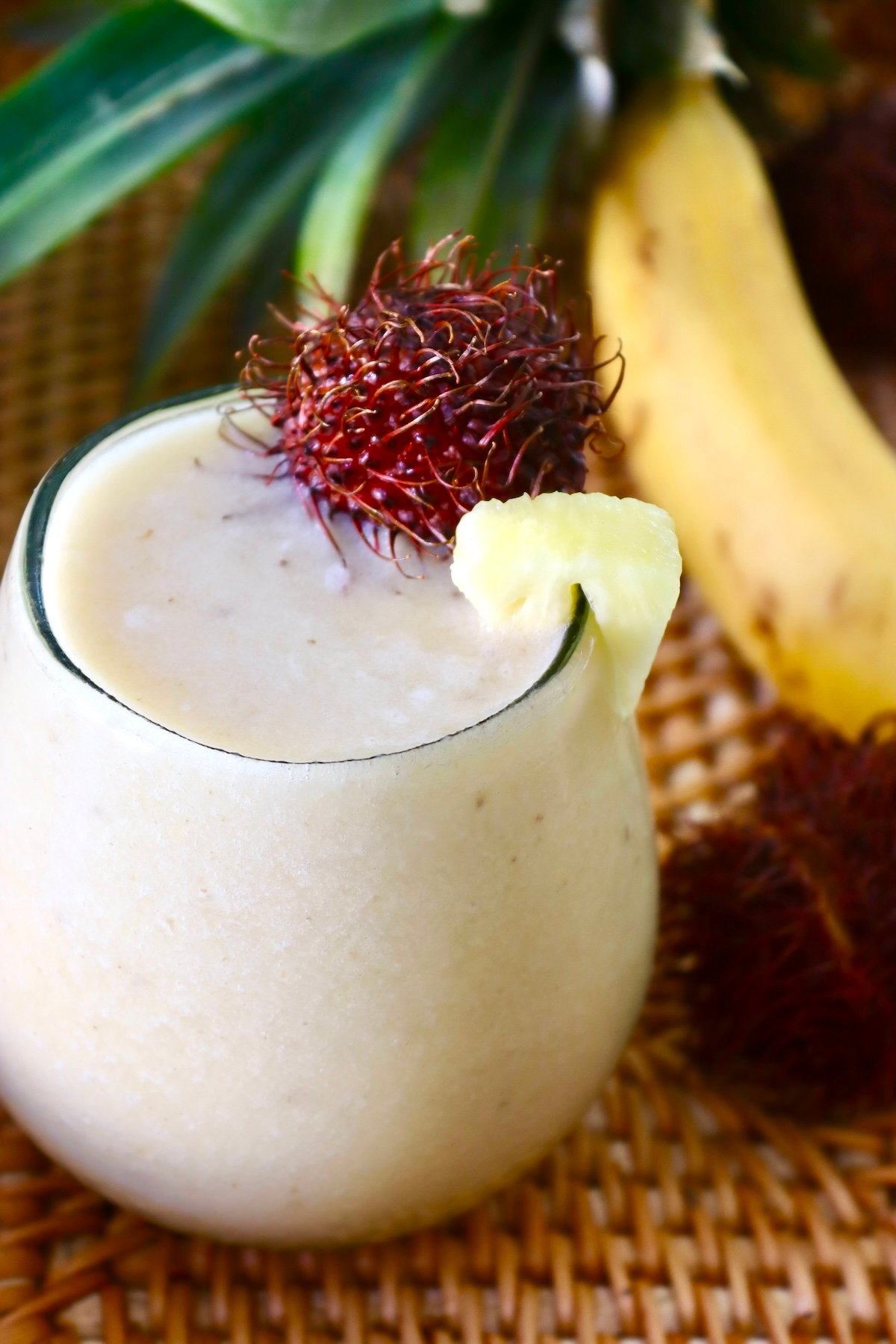 Tropical-Smoothie-Recipe-with-Rambutans Rounded glass filled pale yellow-colored smoothie with a rambutan on the edge.