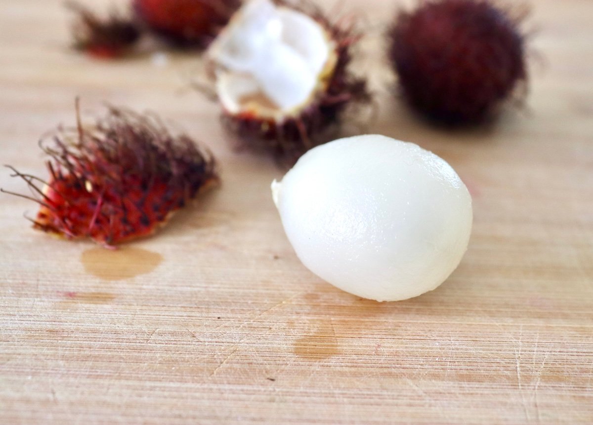 Peeled rambutan with a few rambutan behind it, on light wood cutting board.