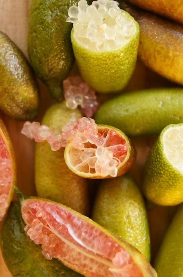 Several Finger Limes on a cutting board, a couple are cut in half with pink "bubbles" of citrus juice spilling out. 