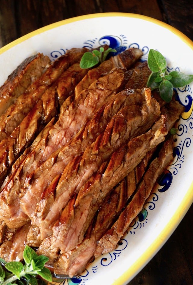 Yellow-rimmed white platter with slices or grilled coffee balsamic flank stea and a couple sprigs of fresh oregano.
