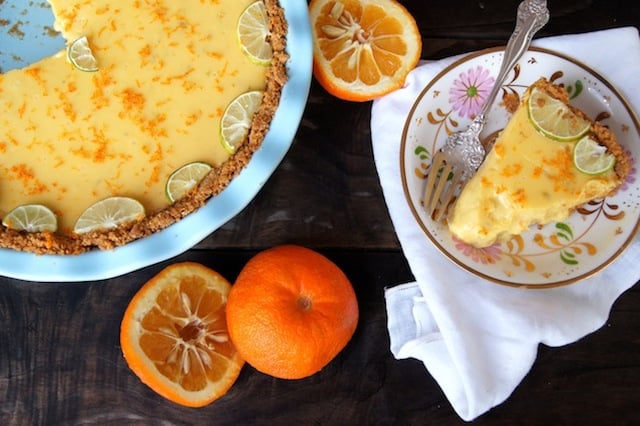 Seville Orange-Key Lime Pie with Cardamom Crust in a light blue pie plate with one slice on a floral plate and a couple of the oranges cut in half