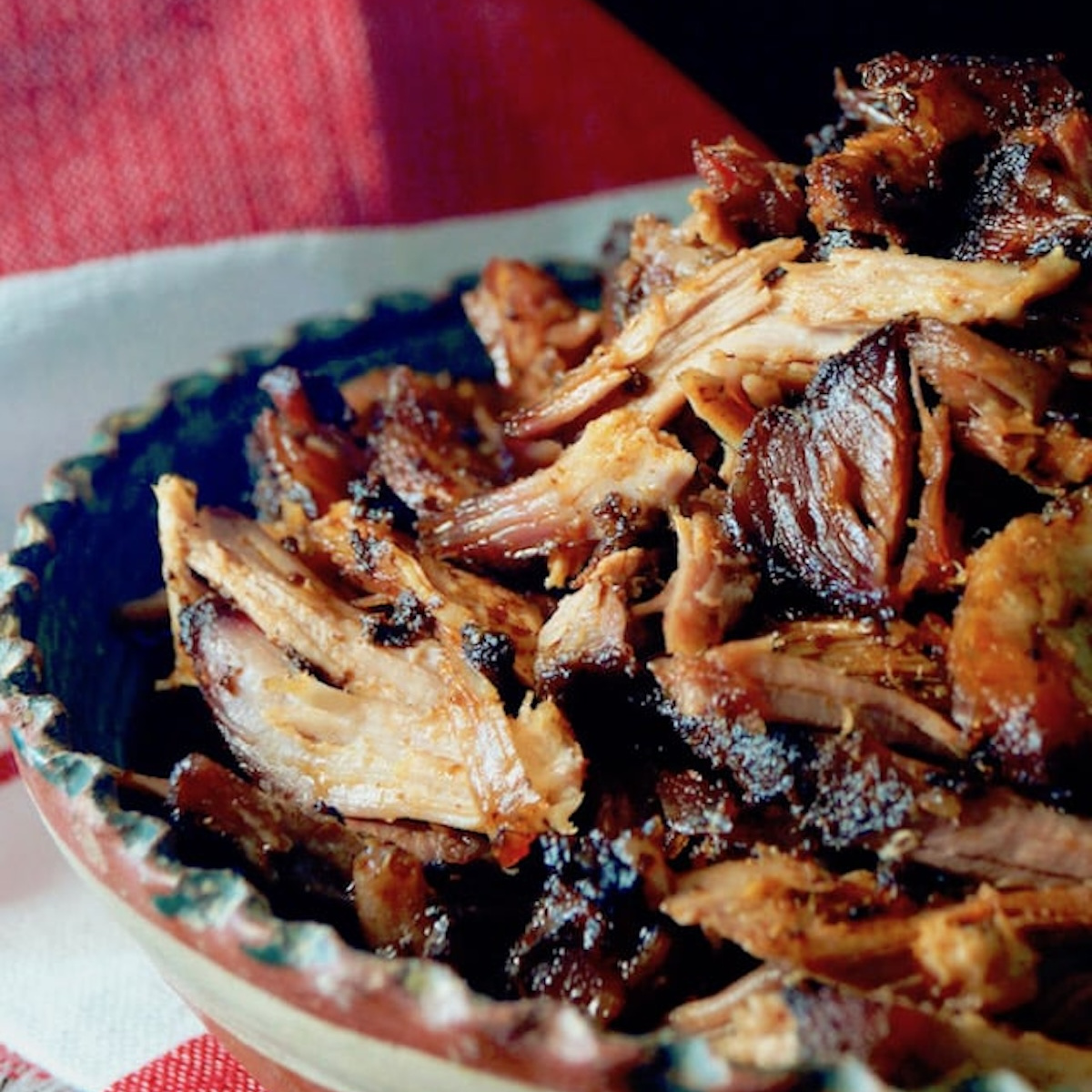 Pickled-Tomato-Pulled-Pork-Tacos Perfect Spiced Pulled Pork, in pieces in a terra cotta bowl with a red and white cloth behind it.