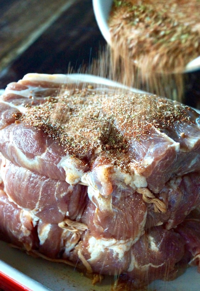 Raw Boston Butt with spice rub being sprinkled on it, for Perfect Spiced Pulled Pork Recipe.