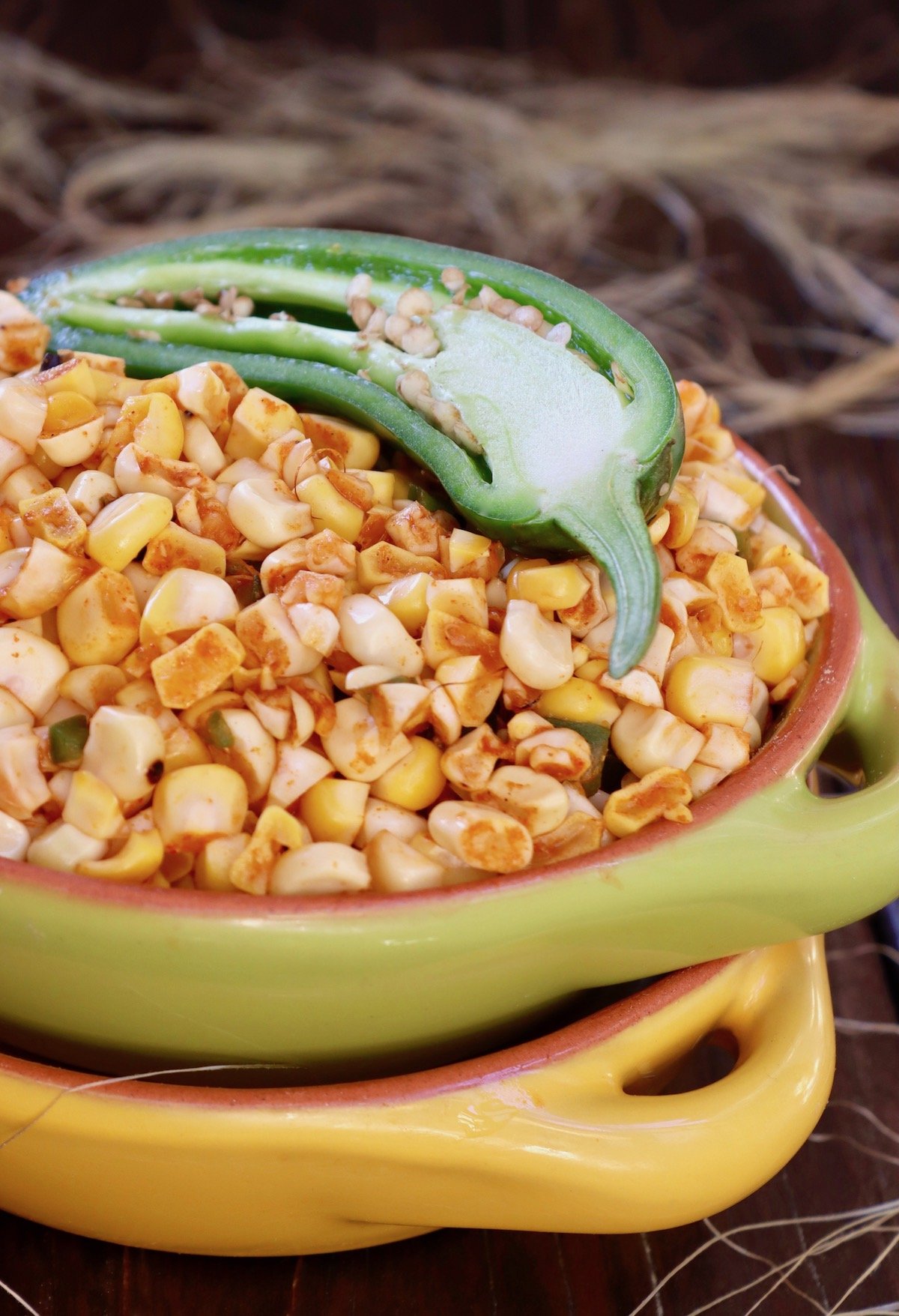 Green bowl filled with fire roasted corn and half of a raw jalapeno on wood.